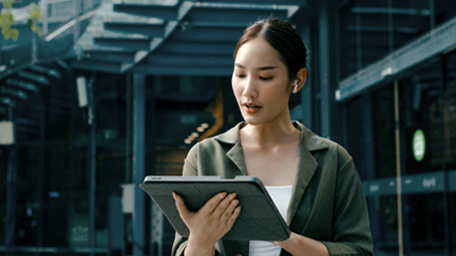 Closeup of young Asian businesswoman discussion walking and using digital tablet and wireless earphones outside modern office building.