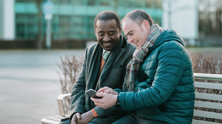 Two businessmen are sitting on a bench in a business park looking at a smart phone together.