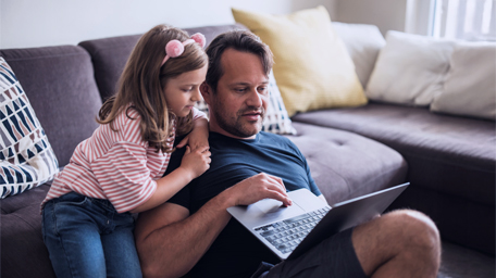 Young girl sitting by her dad using laptop